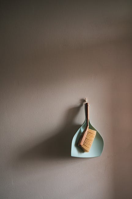 A close-up view of a plain, light-colored wall in an indoor setting, with a small hook holding a green, oval-shaped wall organizer. Inside the organizer, there is a wooden-handled scrub brush with nat